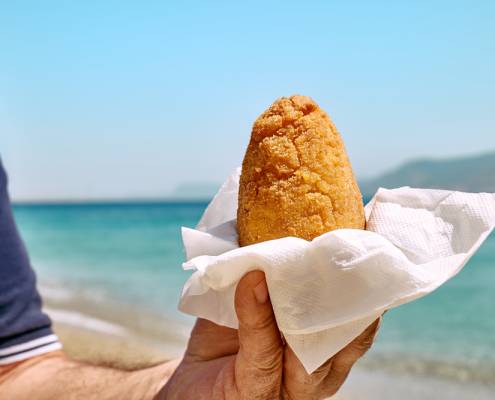Typical Sicilian street food on seaside background. Hand of man holding hot palatable arancina (deep fried rice balls with meat) in the beach. Take out food.