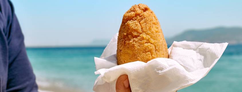 Typical Sicilian street food on seaside background. Hand of man holding hot palatable arancina (deep fried rice balls with meat) in the beach. Take out food.