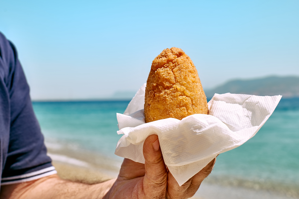 Typical Sicilian street food on seaside background. Hand of man holding hot palatable arancina (deep fried rice balls with meat) in the beach. Take out food.
